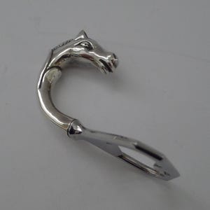 May include: A silver-toned metal bottle opener featuring a sculpted horse head handle. The design includes a curved neck that transitions into a functional flat metal opener end. The surface is polished and reflective, showing clear details of the horse's mane, eyes, and snout in a metallic finish.