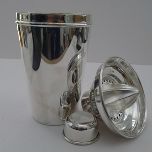 May include: A polished silver cocktail shaker set, comprising a shaker, lid, strainer, and citrus reamer. The set is made of reflective metal. The shaker has a curved design. The set is ideal for making cocktails.