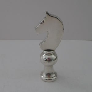May include: A polished silver-toned metal finial featuring a stylized horse head silhouette mounted atop a rounded, tiered pedestal base. The object has a smooth, reflective surface and a solid, upright structure designed for decorative use.