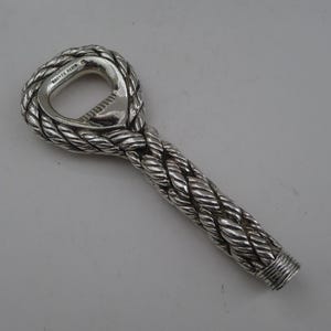 May include: A silver bottle opener with a rope design. The opener has a loop at the top and a handle with a twisted rope pattern. The words "HERMES PARIS" are visible on the top loop.