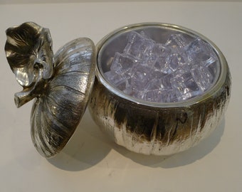 Puede incluir: Hielera plateada con tapa en forma de hoja. La hielera está llena de cubitos de hielo cuadrados y transparentes. La tapa está ligeramente entreabierta, revelando el contenido. La hielera tiene un aspecto texturizado y martillado.