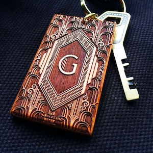 May include: A rectangular wooden keyring with an Art Deco design and the letter "G" in the center. The keyring is brown with intricate carved details and a gold key ring. The word "GRACELAND" is at the bottom.