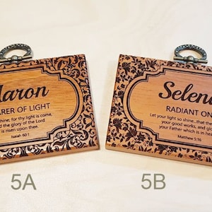 May include: Two wooden plaques with ornate designs and a metal hanging loop. The plaque on the left has the name "Aaron" and the text "Bearer of Light" with a Bible verse. The plaque on the right has the name "Selena" and the text "Radiant One" with a Bible verse.