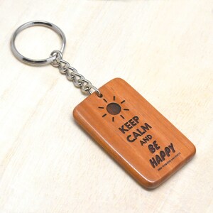Puede incluir: Llavero de madera con un diseño de sol y el texto "Keep Calm and Be Happy".