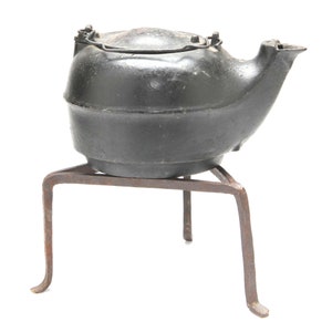 May include: A black cast iron kettle with a lid and a spout, sitting on a three-legged iron stand.