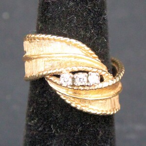 May include: A gold-toned ring with a textured leaf design and three small, clear stones. The ring has a braided rope-like border and is displayed on a black background.