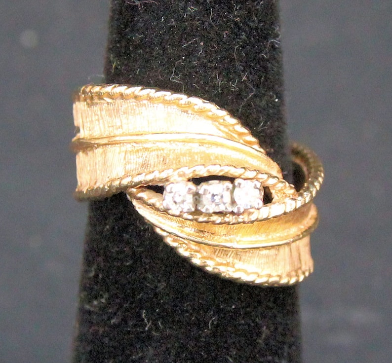 May include: A gold-toned ring with a textured leaf design and three small, clear stones. The ring has a braided rope-like border and is displayed on a black background.