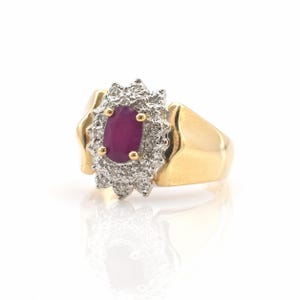 Può includere: Un anello d'oro con un grande rubino ovale circondato da un alone di piccoli diamanti. La fascia dell'anello è in oro lucido e il rubino è di un rosso intenso. L'anello è presentato su uno sfondo bianco.