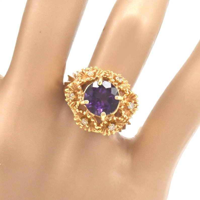 May include: A gold ring with a large, round, deep purple gemstone. The gemstone is set in a decorative, floral-style mount with small, clear accent stones. The ring's band is also gold-toned, creating a striking contrast.
