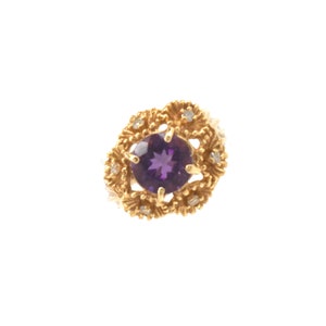 May include: A gold ring with a large, round, deep purple gemstone at the centre, encircled by smaller clear stones and an elaborate gold filigree pattern. The ring is set against a white backdrop.