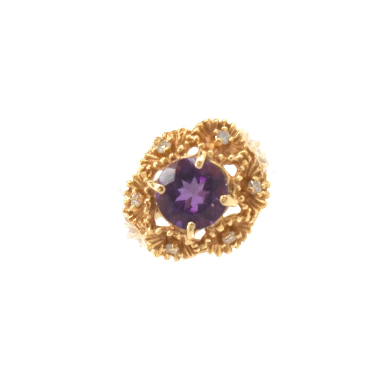 May include: A gold ring with a large, round, deep purple gemstone at the centre, encircled by smaller clear stones and an elaborate gold filigree pattern. The ring is set against a white backdrop.