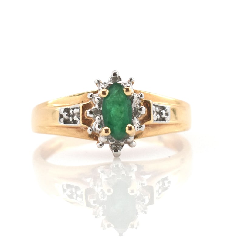 May include: A gold ring featuring a central marquise-cut green gemstone surrounded by a cluster of small clear stones set in white metal. The band is polished yellow gold with additional small clear stones embedded on each side of the setting.