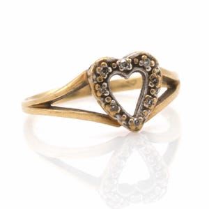 May include: A gold ring featuring a heart-shaped centerpiece. The heart is outlined in a silver-toned metal and is set with small, clear, round stones along its perimeter. The band of the ring splits into two sections as it reaches the heart.
