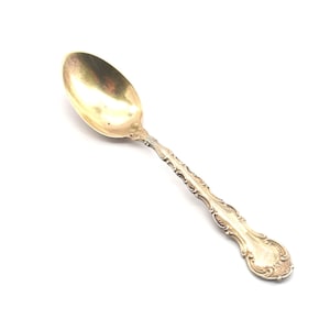 May include: A gold-toned teaspoon with an ornate, detailed handle. The spoon has a rounded bowl and a classic design.