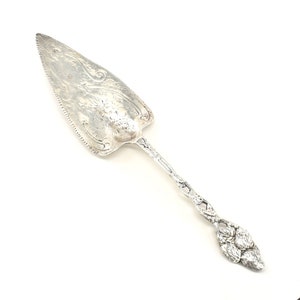 May include: A silver cake server with an ornate, floral design. The handle is shaped like a cluster of grapes.