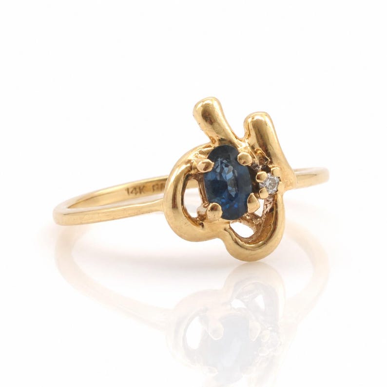 May include: A 14K gold ring featuring an oval-cut blue sapphire gemstone and a small round-cut white diamond. The band has a polished finish and the metal is stamped with 14K on the inner surface. The setting has an asymmetrical, curved design.