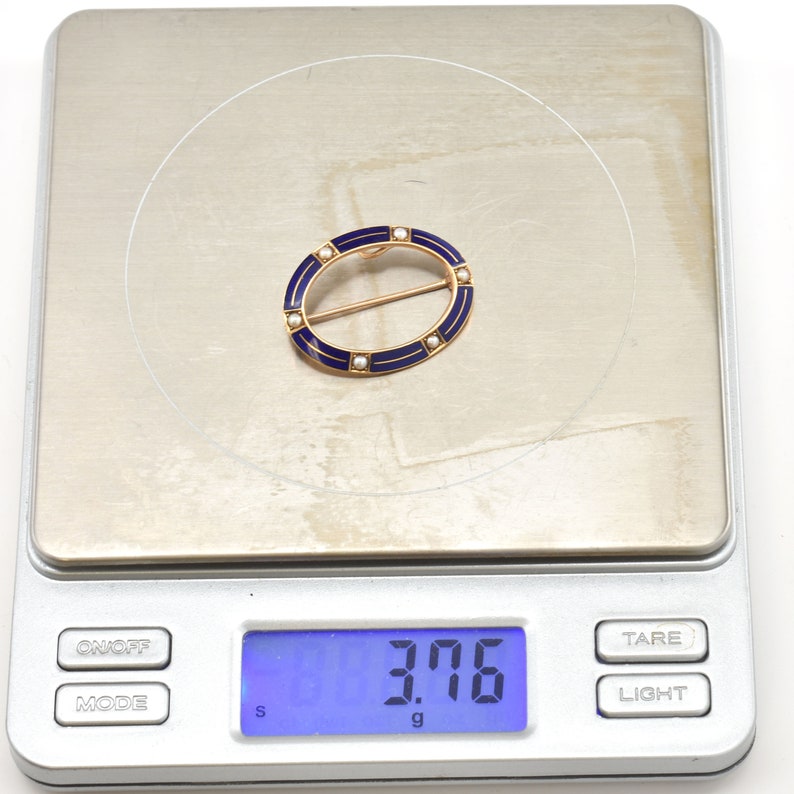 May include: A gold oval brooch with blue enamel and white pearl accents. The brooch weighs 3.76 grams.