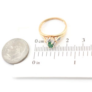 May include: A gold ring featuring an oval-cut green emerald center stone surrounded by small clear accent stones. The ring is positioned next to a U.S. dime and a ruler showing a width of approximately 0.5 inches for the stone setting.