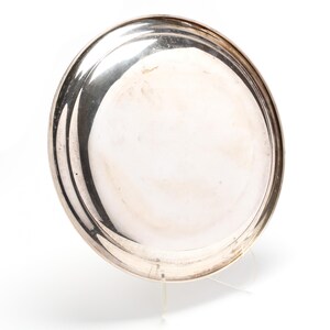 May include: A round, silver-colored metal tray with a slightly raised rim. The tray has a reflective surface and appears to be made of polished metal. The tray is displayed on a clear stand against a white background.