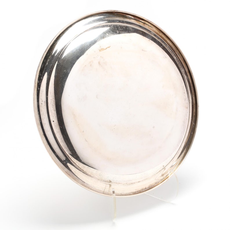 May include: A round, silver-colored metal tray with a slightly raised rim. The tray has a reflective surface and appears to be made of polished metal. The tray is displayed on a clear stand against a white background.