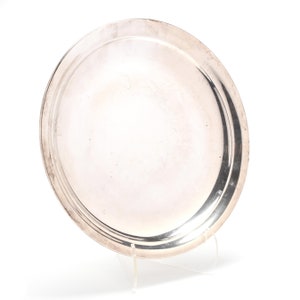 May include: A round, silver-colored serving tray with a raised rim. The tray is displayed on a clear acrylic stand against a white background. The reflective surface of the tray shows slight imperfections.