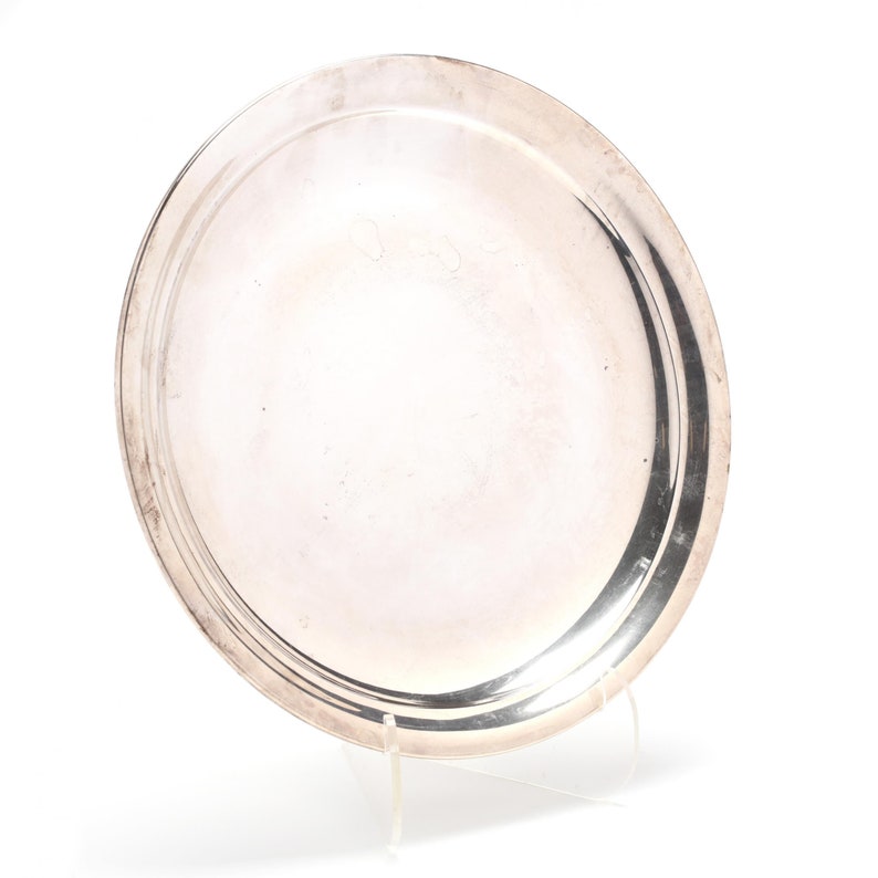 May include: A round, silver-colored serving tray with a raised rim. The tray is displayed on a clear acrylic stand against a white background. The reflective surface of the tray shows slight imperfections.