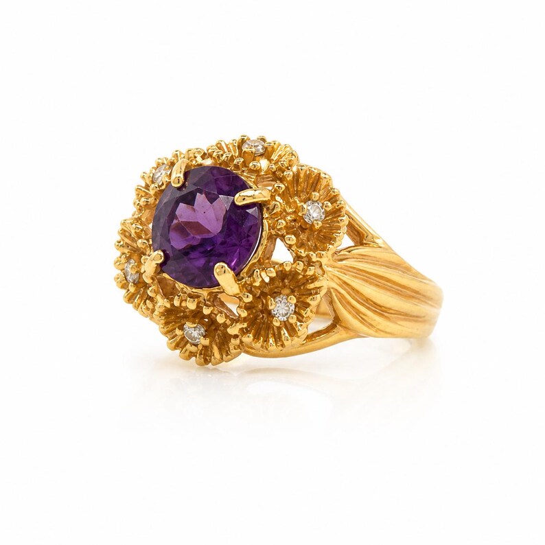May include: A gold ring with a large, oval-cut purple amethyst gemstone, surrounded by small diamonds and a floral design. The ring's band has a textured, ribbed pattern, and the setting is also gold.