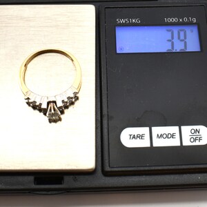 May include: A gold ring with a cluster of small diamonds and a larger teardrop-shaped diamond. The ring is on a digital scale, weighing 3.9 grams.