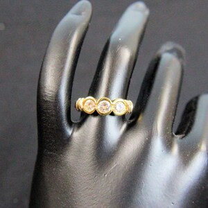 May include: A gold ring featuring three round, clear, faceted stones set in a horizontal row. The stones are held in place by individual circular gold bezels, creating a continuous band design that sits across the finger with a polished finish.