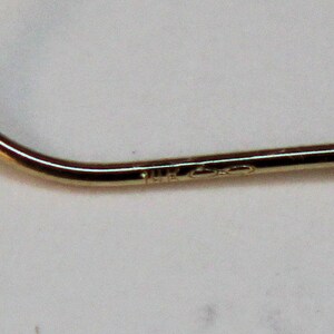 K&ouml;nnte beinhalten: Ein gebogenes Golddrahtelement, wahrscheinlich Teil eines Ohrrings, mit einer glatten, polierten Metalloberfl&auml;che. Das Metall ist mit dem Text 14K gepr&auml;gt, was den Goldgehalt angibt, und ein Unendlichkeitssymbol ist neben dem Stempel sichtbar.