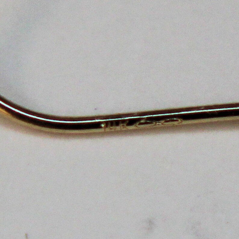 K&ouml;nnte beinhalten: Ein gebogenes Golddrahtelement, wahrscheinlich Teil eines Ohrrings, mit einer glatten, polierten Metalloberfl&auml;che. Das Metall ist mit dem Text 14K gepr&auml;gt, was den Goldgehalt angibt, und ein Unendlichkeitssymbol ist neben dem Stempel sichtbar.