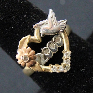 Peut inclure: Bague bicolore or et argent en forme de cœur. La bague présente une colombe, une fleur et le chiffre "2000". De petites pierres claires sont serties sur le côté droit du cœur. Bijou de style vintage.
