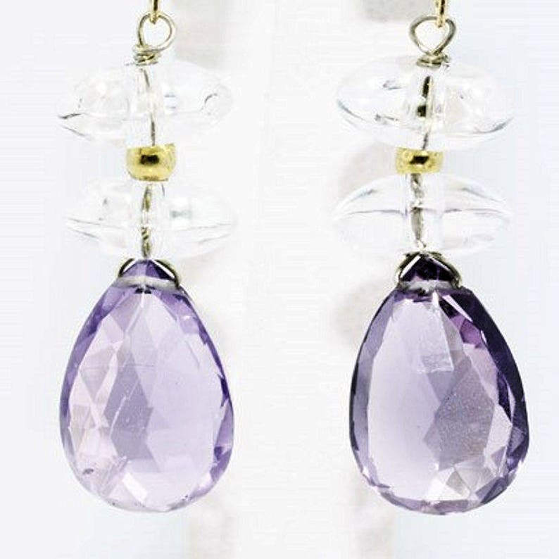 K&ouml;nnte beinhalten: Ein Paar Ohrh&auml;nger mit facettierten, birnenf&ouml;rmigen violetten Amethyststeinen, die an zwei klaren, runden, scheibenf&ouml;rmigen Perlen und einer kleinen goldfarbenen Metallperle h&auml;ngen. Die Elemente sind durch d&uuml;nne goldfarbene Metalldr&auml;hte verbunden.