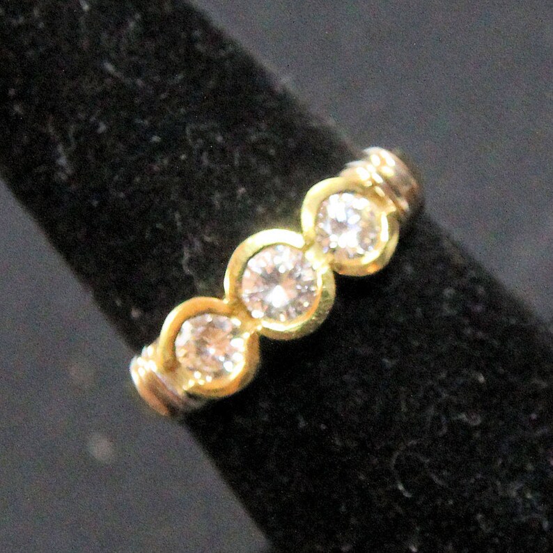 May include: A gold ring featuring three round, clear, faceted stones set in a row. The stones are held in place by individual gold bezels, creating a continuous band design. The metal has a polished finish that reflects light across the surface.