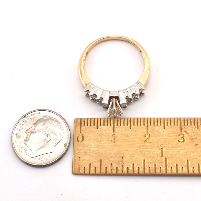 May include: A gold ring with a central diamond and smaller accent stones. The ring is set against a white background with a ruler showing measurements in inches and a dime for size comparison. The ring's design features a cluster of stones.