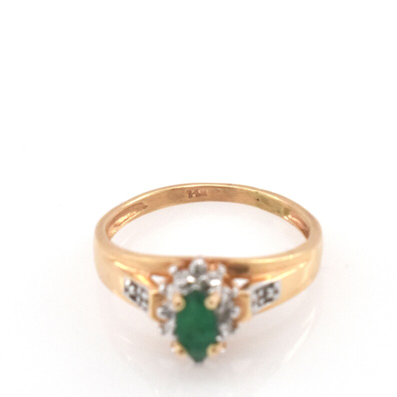 May include: A gold ring featuring a central marquise-cut green gemstone surrounded by a halo of small clear stones. The band is polished gold and includes a clear 14K stamp on the inner surface, indicating the metal purity of the jewelry piece.