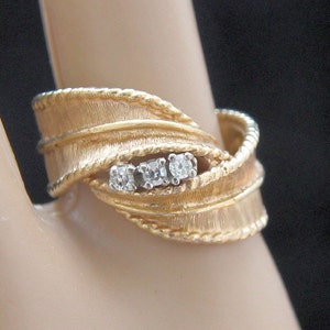 May include: A gold-toned ring with a textured, leaf-like design. The ring features three small, clear gemstones set in a row. The band has a twisted rope-like detail. The ring is displayed on a finger.
