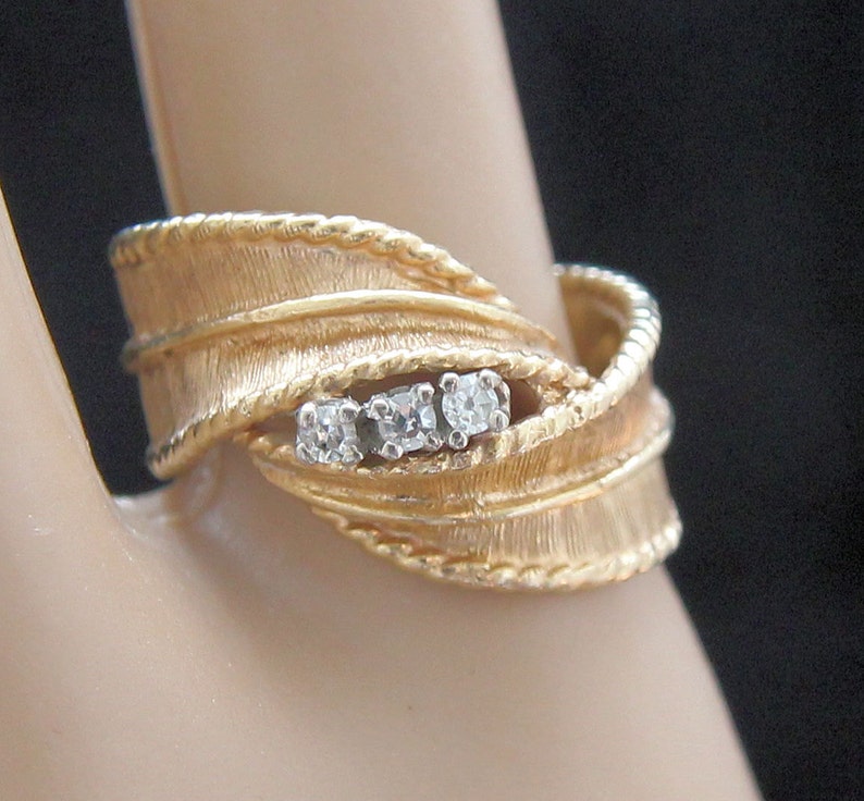 May include: A gold-toned ring with a textured, leaf-like design. The ring features three small, clear gemstones set in a row. The band has a twisted rope-like detail. The ring is displayed on a finger.