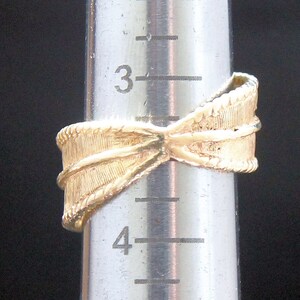May include: A gold-colored ring with a textured bow design. The ring is on a silver measuring tool. The ring's design includes a textured band and a detailed bow shape, showcasing a classic and elegant style.