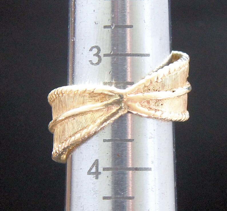 May include: A gold-colored ring with a textured bow design. The ring is on a silver measuring tool. The ring's design includes a textured band and a detailed bow shape, showcasing a classic and elegant style.