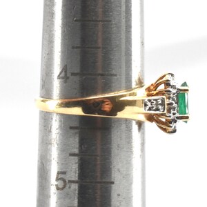 May include: A gold ring featuring a rectangular green gemstone surrounded by small clear stones, set on a metal ring mandrel showing a size between 4 and 5 inches. The band is polished gold, and the setting includes silver-toned metal accents.