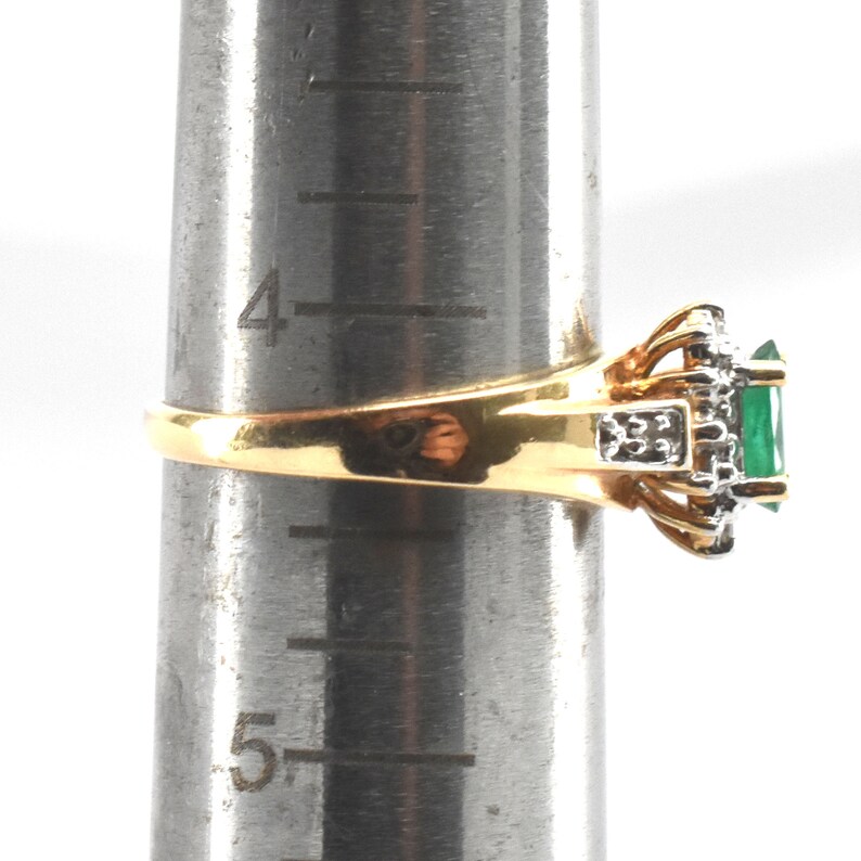May include: A gold ring featuring a rectangular green gemstone surrounded by small clear stones, set on a metal ring mandrel showing a size between 4 and 5 inches. The band is polished gold, and the setting includes silver-toned metal accents.
