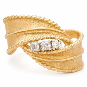 May include: A gold-toned ring with a leaf-like design. The ring features three small, clear gemstones set in the center. The band has a textured finish and a rope-like detail around the edges, creating an elegant and unique look.