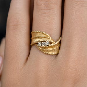 May include: A gold ring with a textured leaf design, featuring three small diamonds. The ring has a twisted rope-like border and is shown on a person's finger. The ring is a piece of jewelry.