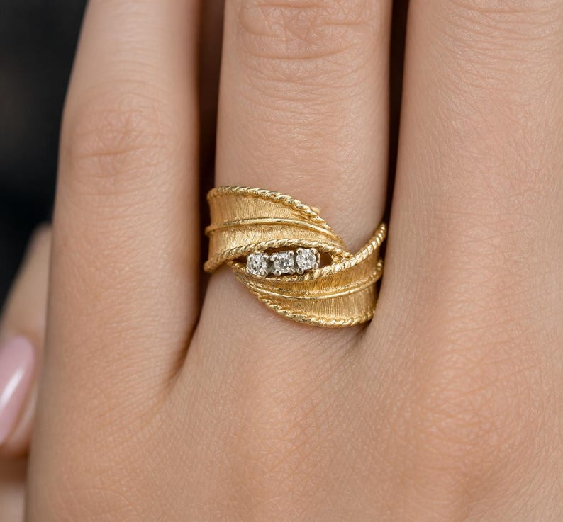 May include: A gold ring with a textured leaf design, featuring three small diamonds. The ring has a twisted rope-like border and is shown on a person's finger. The ring is a piece of jewelry.