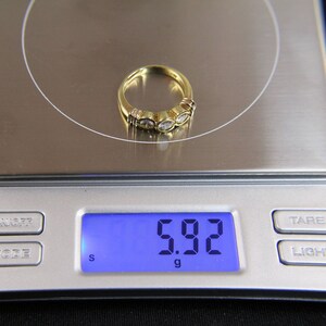 May include: A gold ring featuring three round, clear stones set in a row, resting on a digital scale that displays a weight of 5.92 grams. The band is polished yellow gold, and the stones are held in place by individual metal settings.