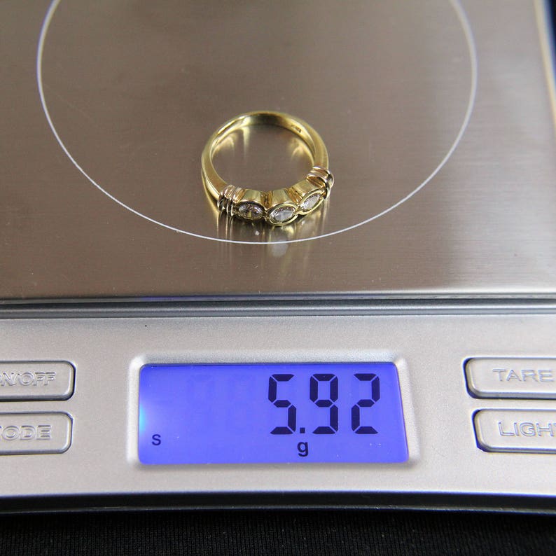May include: A gold ring featuring three round, clear stones set in a row, resting on a digital scale that displays a weight of 5.92 grams. The band is polished yellow gold, and the stones are held in place by individual metal settings.