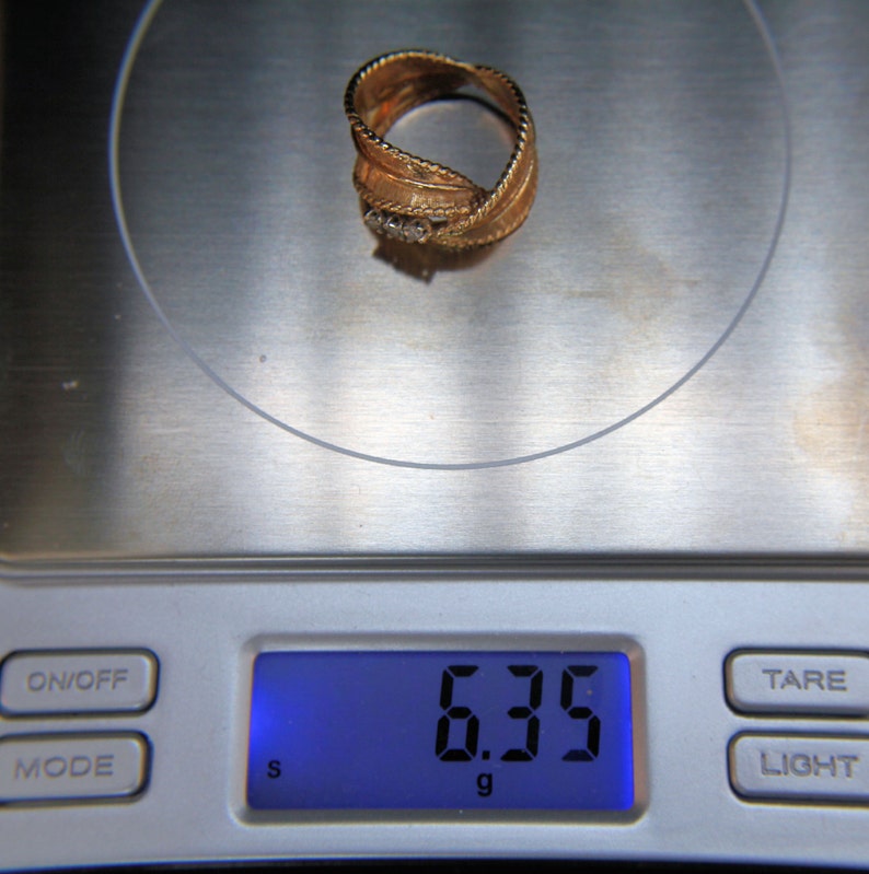 May include: A gold-colored ring with a textured design and three small, clear stones. The ring is on a digital scale, which displays a weight of 6.35 grams.