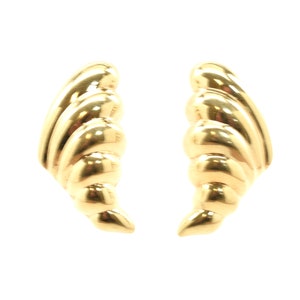 May include: A pair of gold-coloured earrings. Each earring features a curved, shell-like design with a series of rounded ridges. The earrings are set against a white background, highlighting their polished, reflective surface.