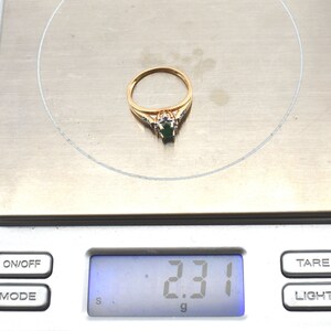 May include: A gold ring featuring a central oval-cut green gemstone surrounded by small clear stones, resting on a digital scale that displays a weight of 2.31 grams. The band is thin and polished, showcasing a classic design with a prong setting.